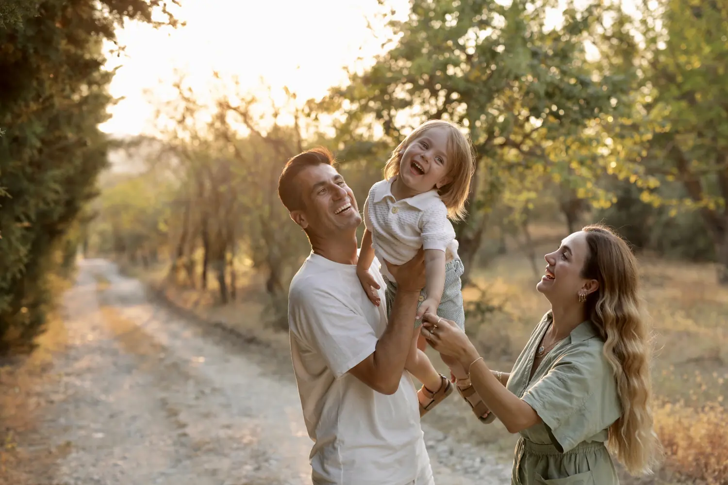 Oğuzhan lifting their laughing son into the air while Fatma reaches up towards him, on a tree-lined path in warm evening light.
